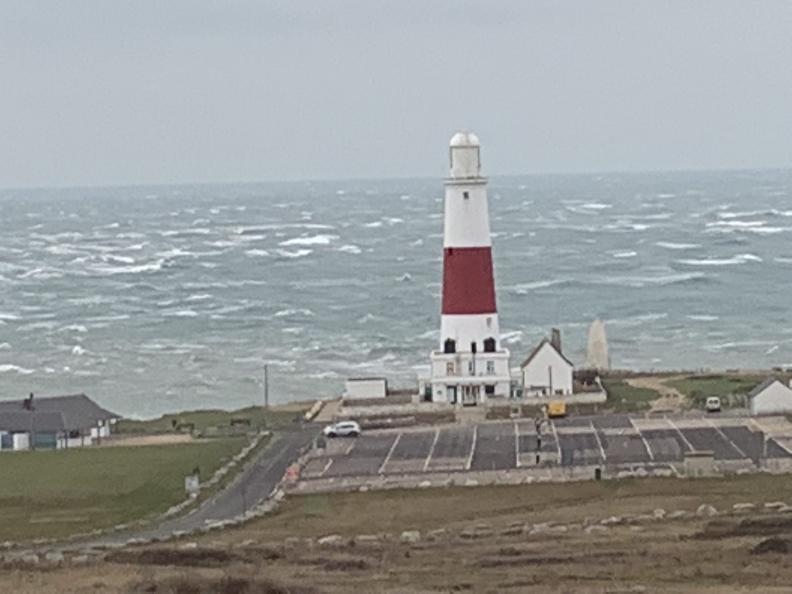 A lighthouse with a red and white striped tower stands near the ocean. The sea is choppy with white waves. Surrounding the lighthouse are a few small buildings and a parking area. The sky is overcast, and the landscape is grassy and open.