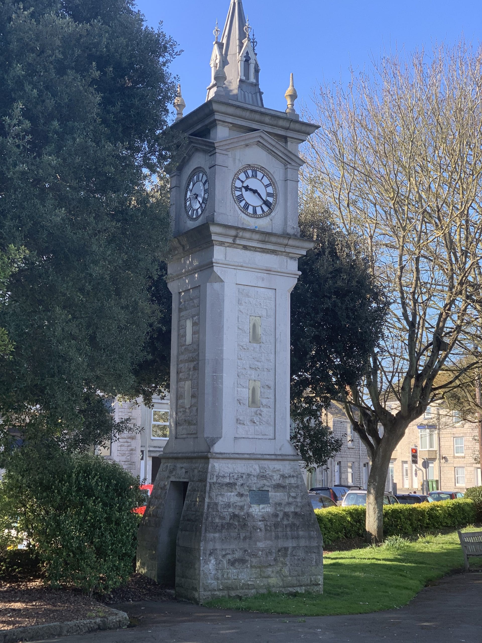 A tall, stone clock tower stands amidst greenery on a sunny day. Its four clock faces show the same time, and the tower is surrounded by trees and grass, with a clear blue sky above.