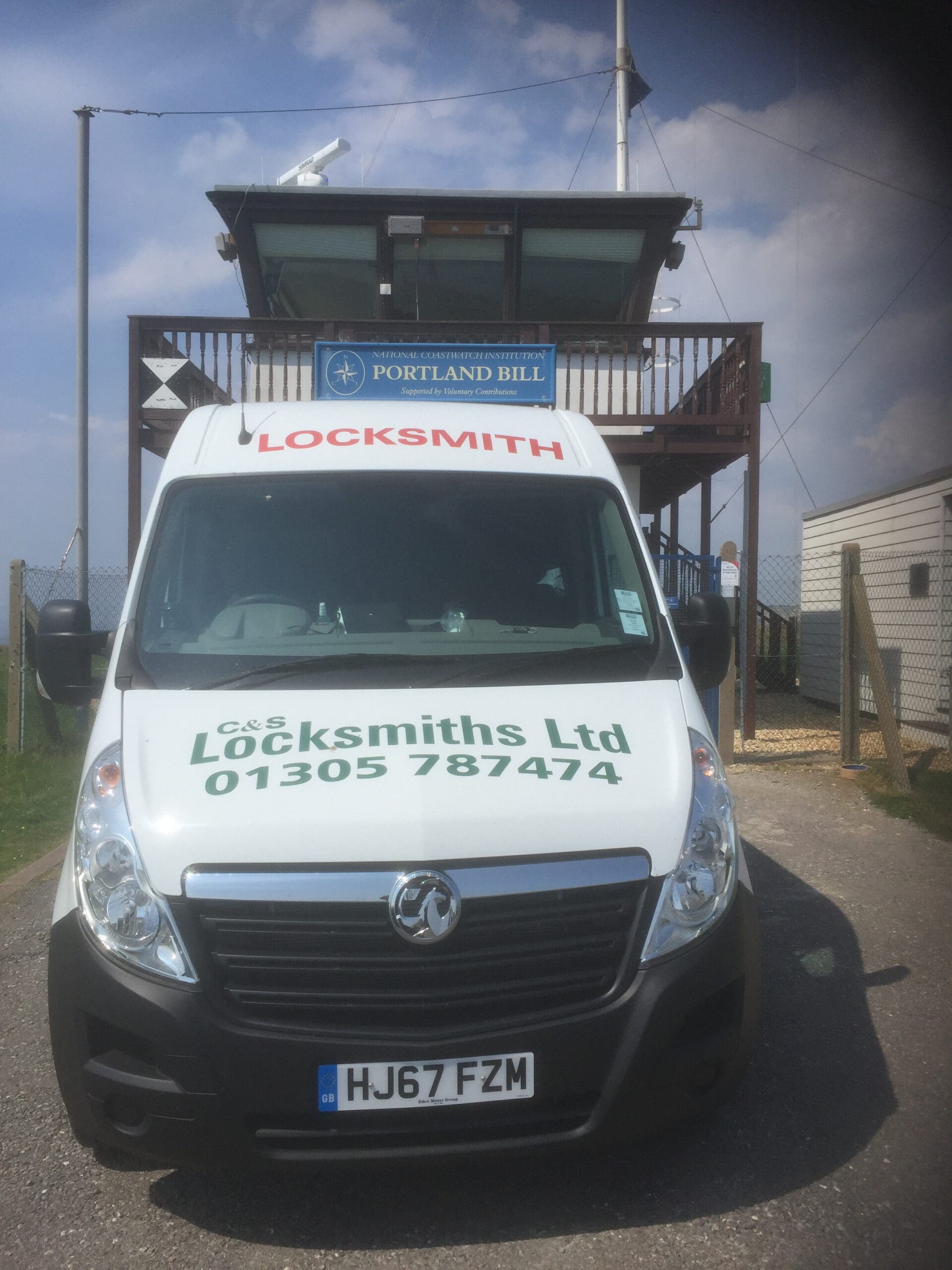 A white locksmith van with signage "C&S Locksmiths Ltd" and a phone number is parked in front of a building with a "Portland Bill" sign. The sky is clear and sunny.