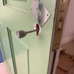 A green door with a removed handle reveals a hole and exposed internal lock mechanism. The latch plate is visible on the edge. Some scratches and chipped paint are evident around the handle area. Cardboard is scattered on the floor in the background.