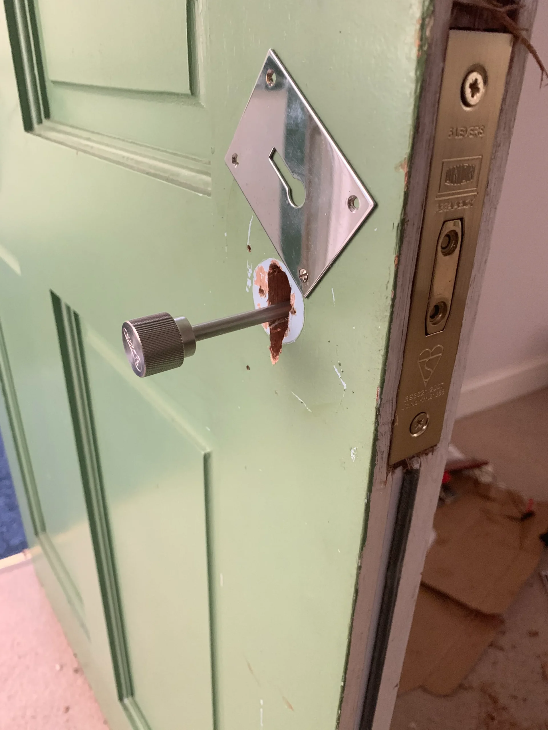 A green door with a removed handle reveals a hole and exposed internal lock mechanism. The latch plate is visible on the edge. Some scratches and chipped paint are evident around the handle area. Cardboard is scattered on the floor in the background.