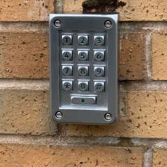 A metallic keypad with numbers 0-9, asterisk, and hashtag is mounted on a brick wall. The keypad has rectangular buttons and is used for secure entry access. Small patches of moss are visible above the keypad on the bricks.