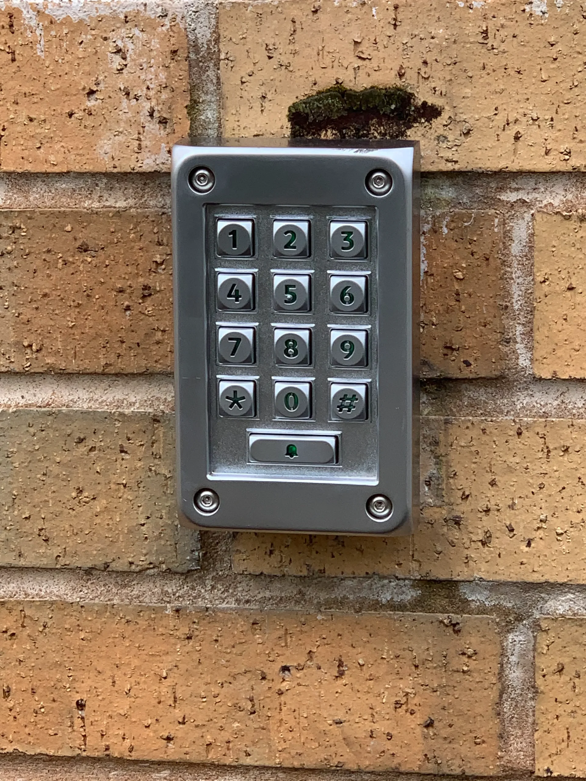 A metallic keypad with numbers 0-9, asterisk, and hashtag is mounted on a brick wall. The keypad has rectangular buttons and is used for secure entry access. Small patches of moss are visible above the keypad on the bricks.