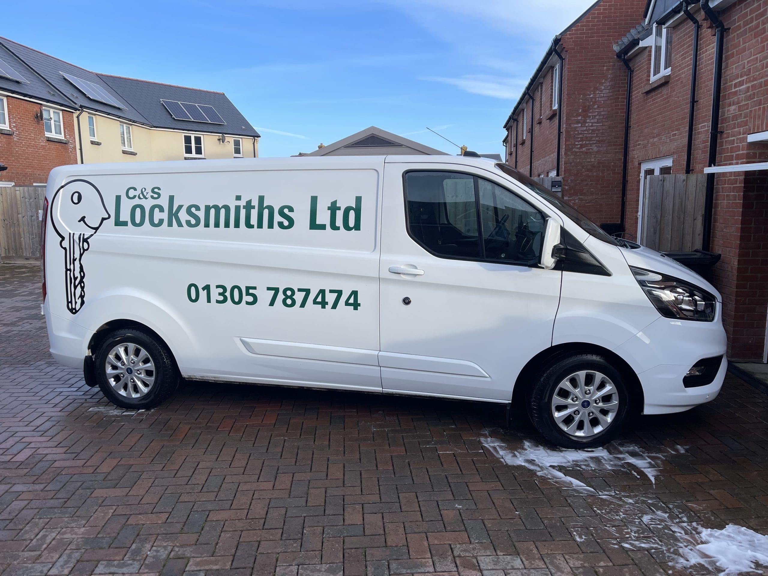 A white locksmith van with "C&S Locksmiths Ltd" and a key logo on the side. The phone number 01305 787474 is displayed. The vehicle is parked on a brick driveway next to residential buildings.