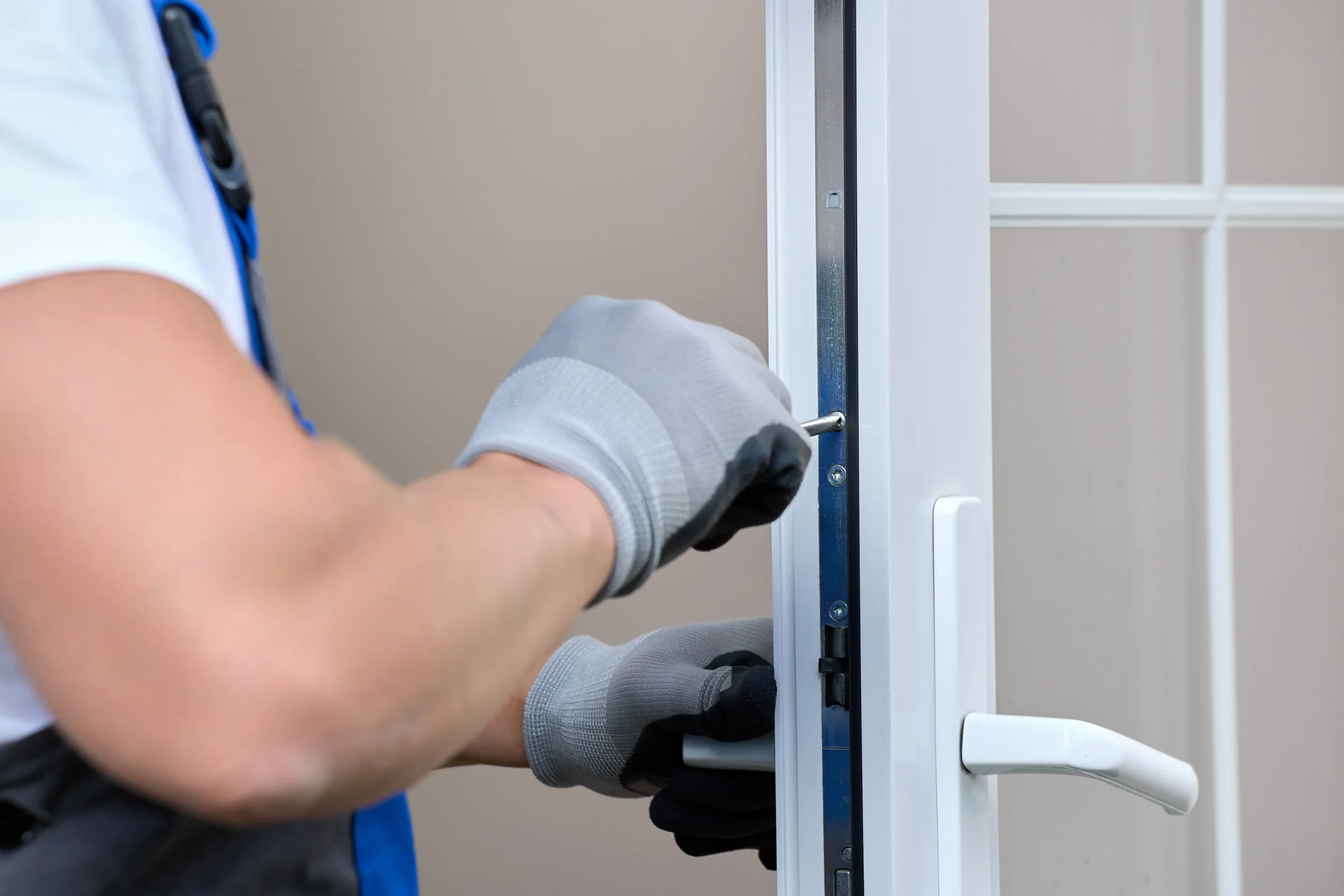 A person wearing gloves is adjusting or repairing the lock mechanism on a white door using a screwdriver. The focus is on the hands and the tool, with the background slightly blurred.