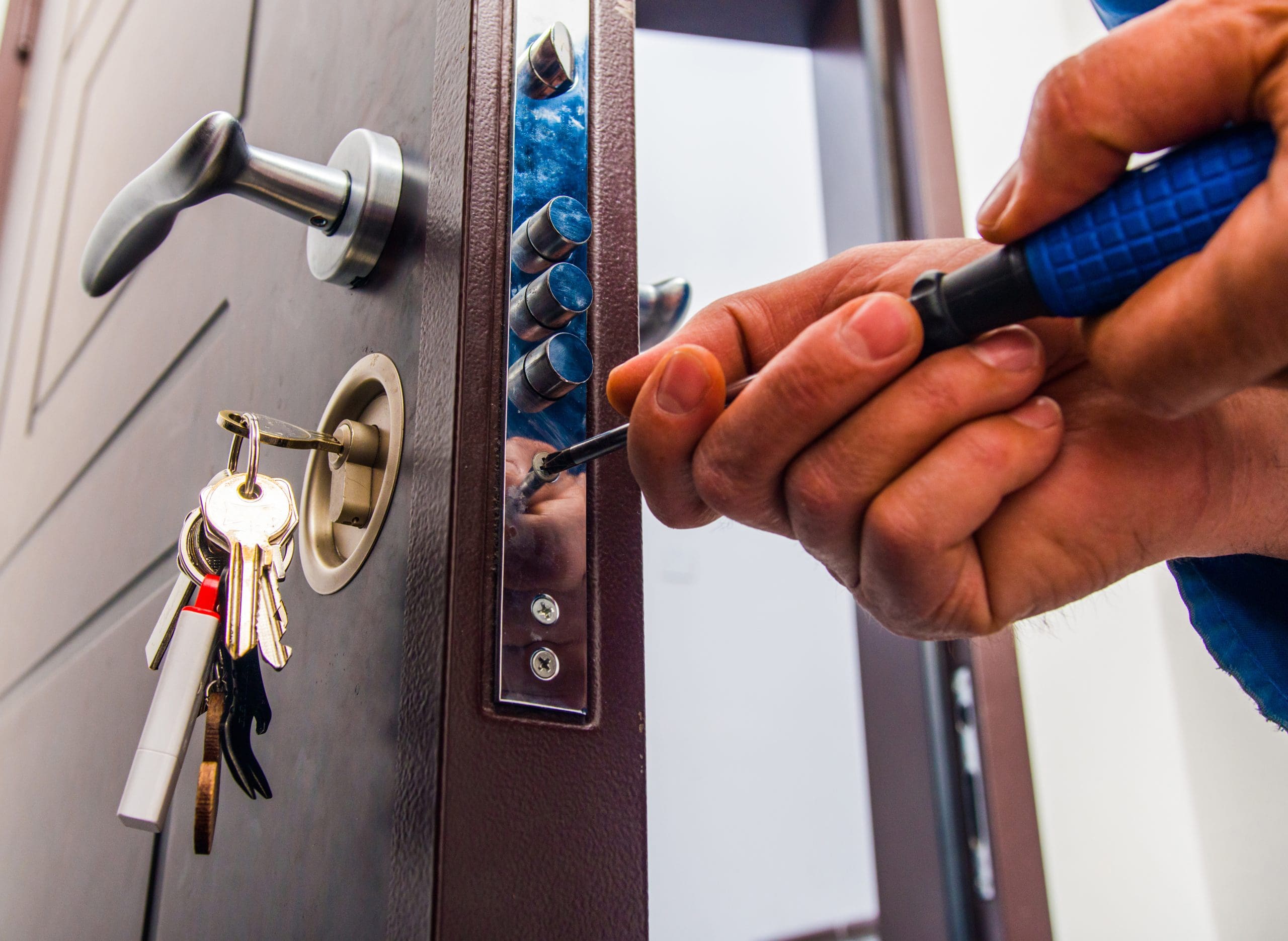 A person repairs a door lock with a screwdriver, focusing on the lock mechanism. A keyring with several keys hangs from the door handle. The door is dark brown with a modern design.