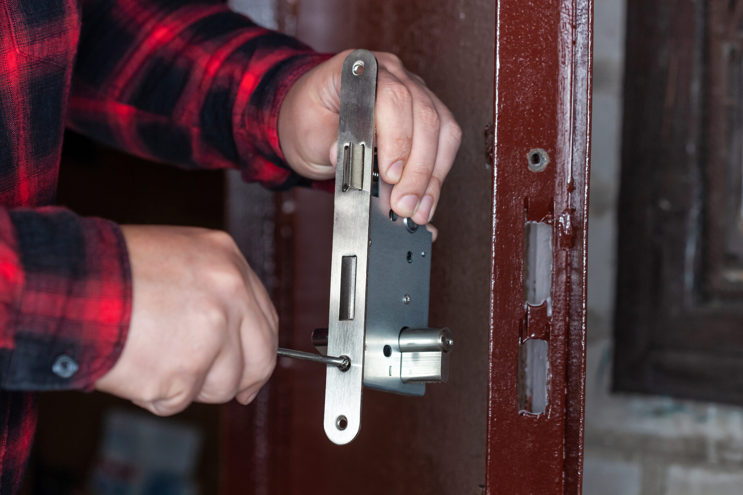 A person in a red plaid shirt installs or repairs a metal door lock using a screwdriver on a brown wooden door.