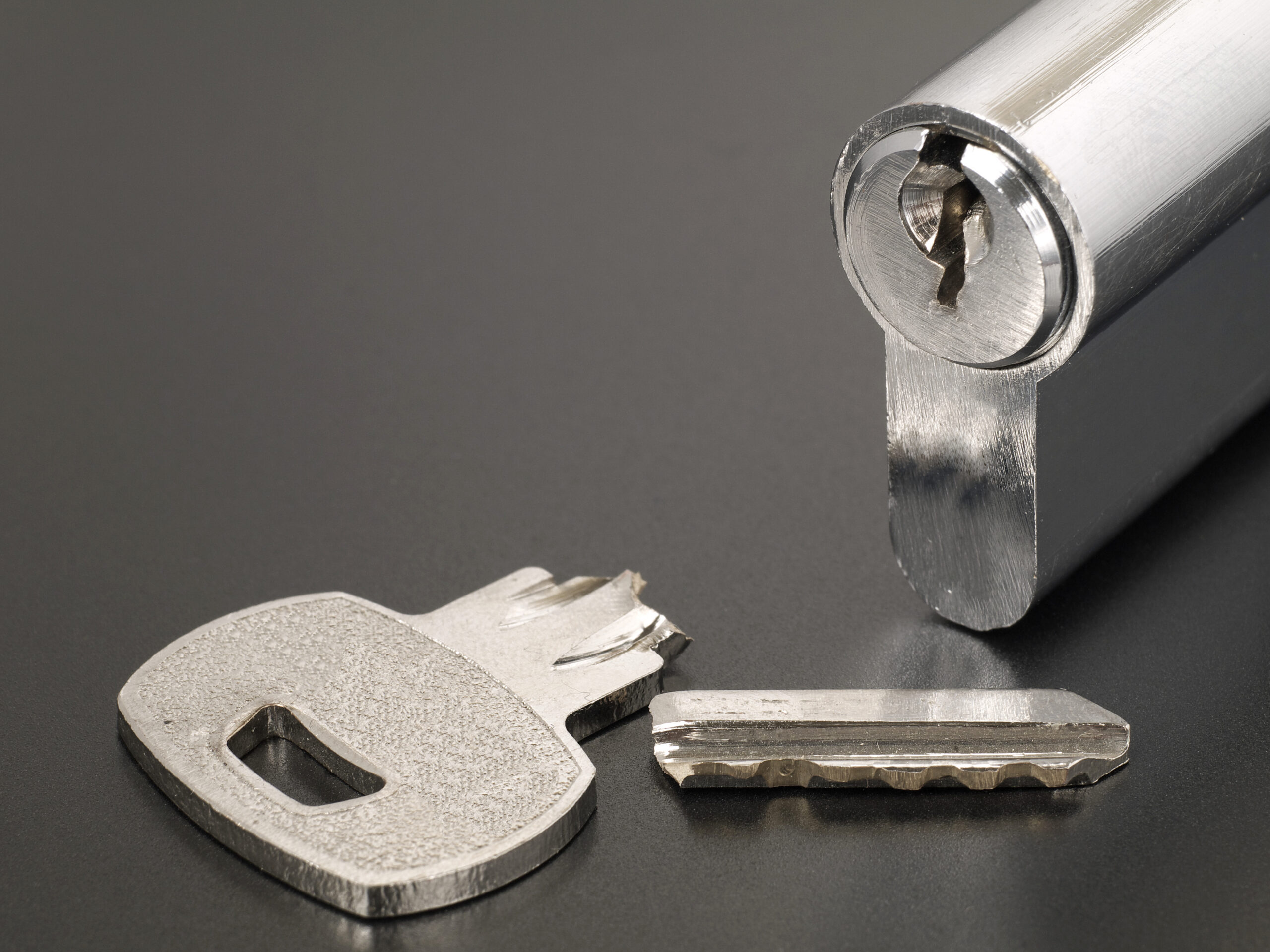 A close-up of a broken key, split into two pieces, lying next to a silver lock cylinder on a dark surface.