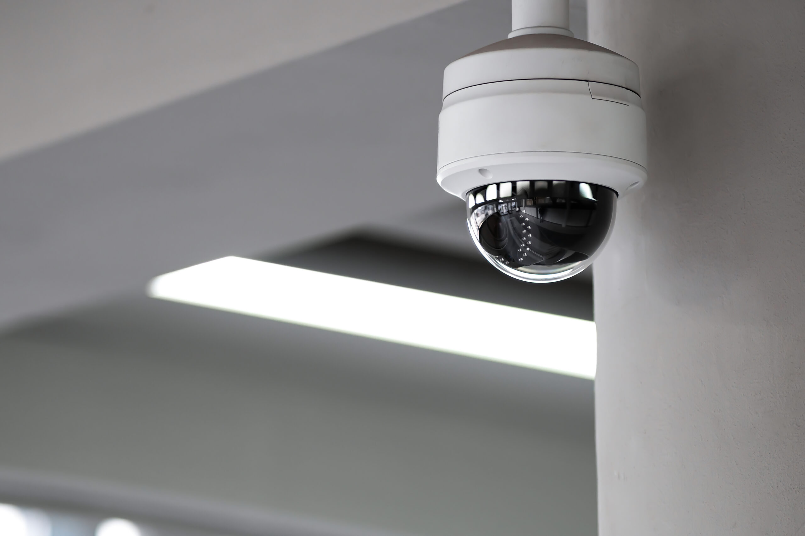 A white dome security camera is mounted on a light-colored indoor wall near the ceiling, with fluorescent lights and structural beams visible in the background.