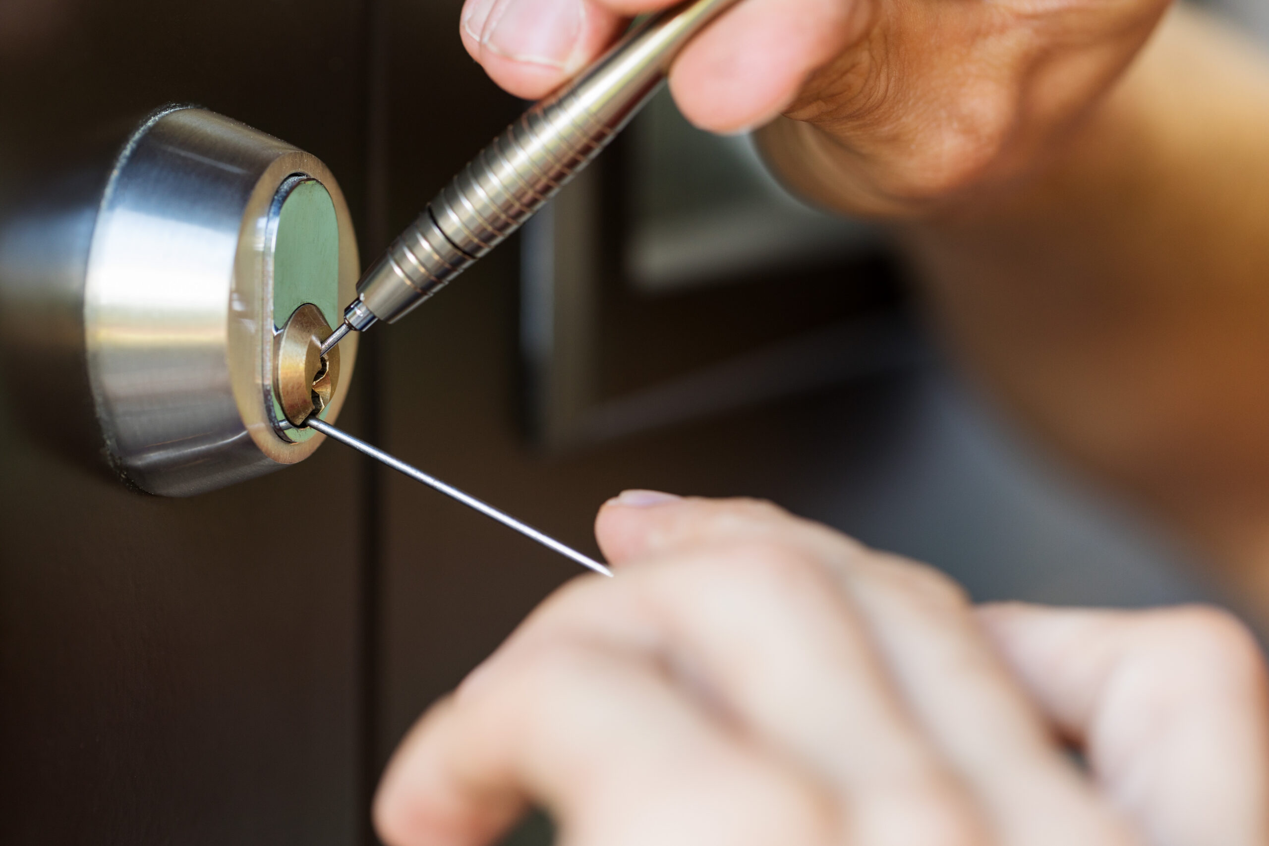 Close-up of hands using lock picking tools to open a door lock, with one tool inserted into the keyhole and another hand holding a tension wrench.