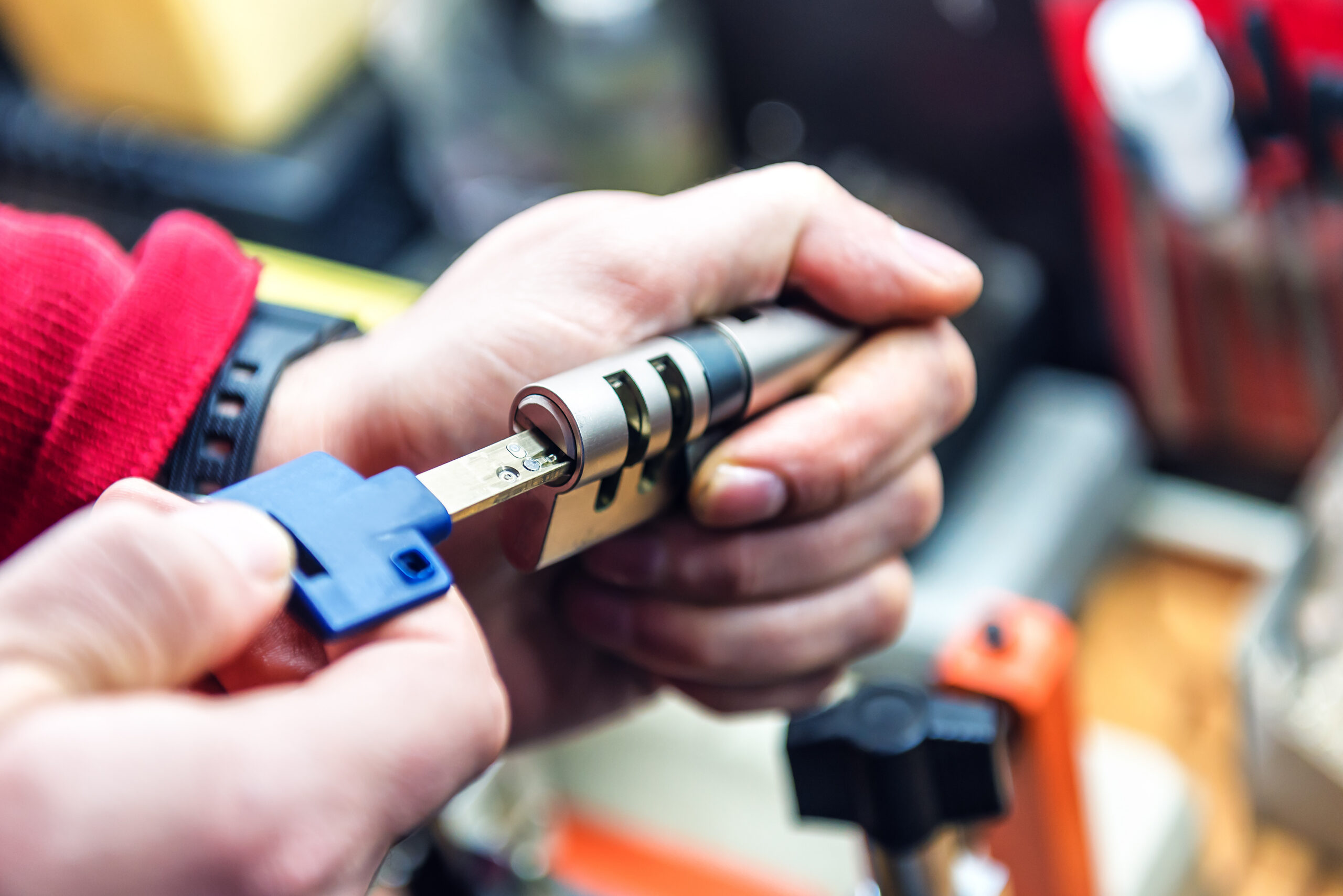 A person’s hands working with a door lock cylinder and a blue-handled key, possibly performing lock repair or rekeying, with tools and equipment visible in the background.