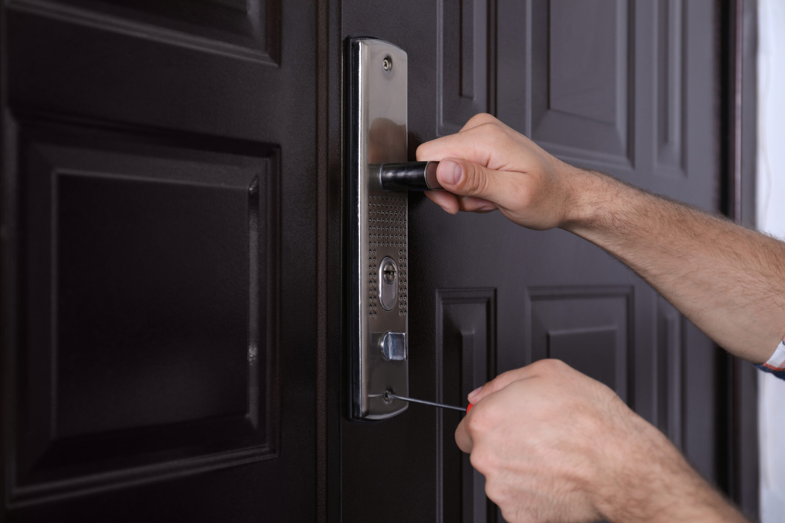 A person uses tools to pick the lock of a dark brown door, with one hand turning the door handle and the other inserting a tool into the lock mechanism.