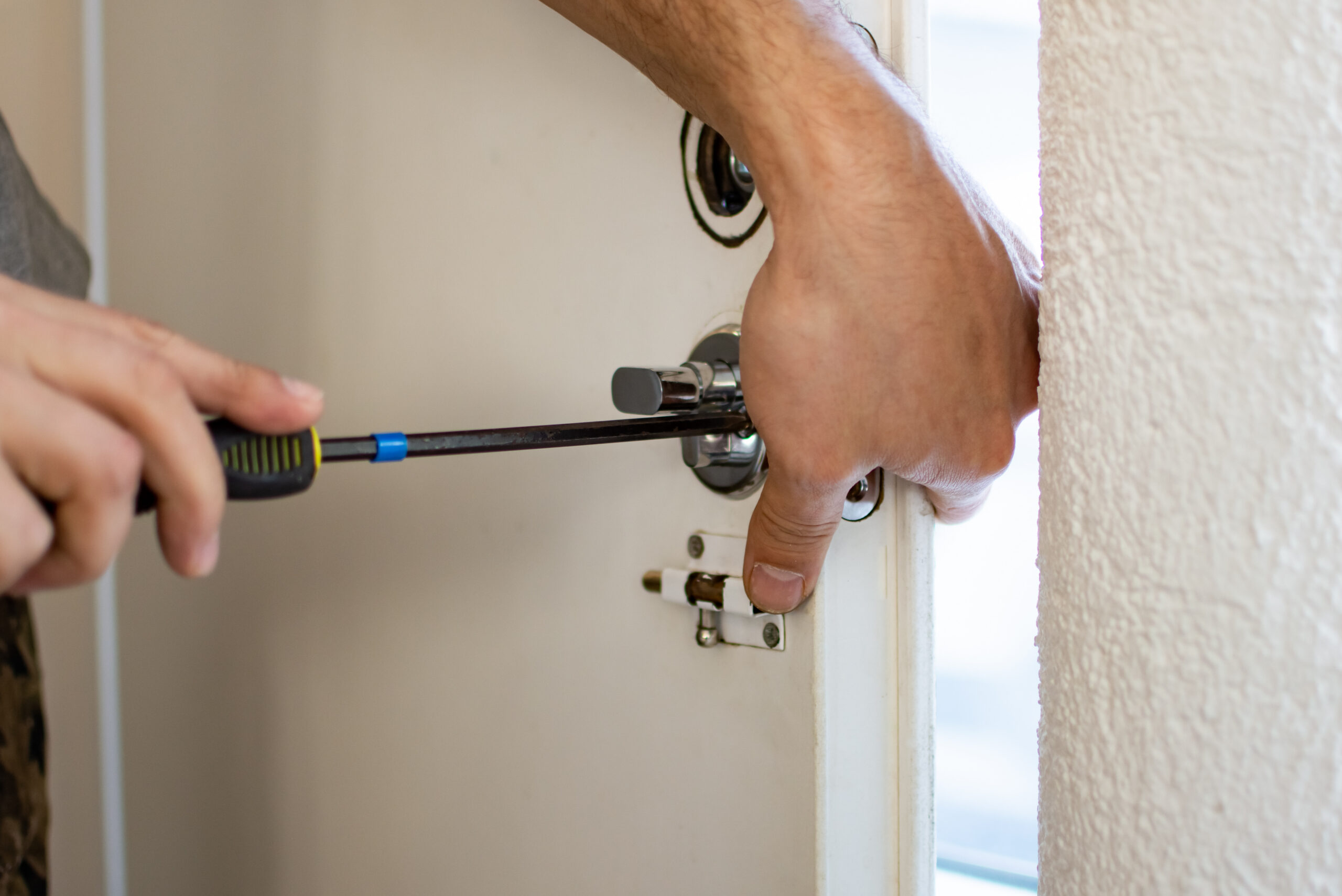 A close-up of a person’s hands using a screwdriver to install or repair a door lock on a white door. The door is slightly open and the focus is on the lock mechanism.