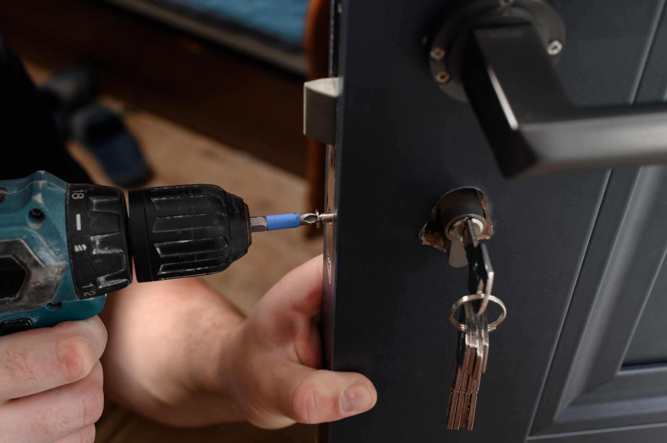 A person uses a cordless power drill to unscrew a screw from a door lock while a set of keys hangs from the lock.