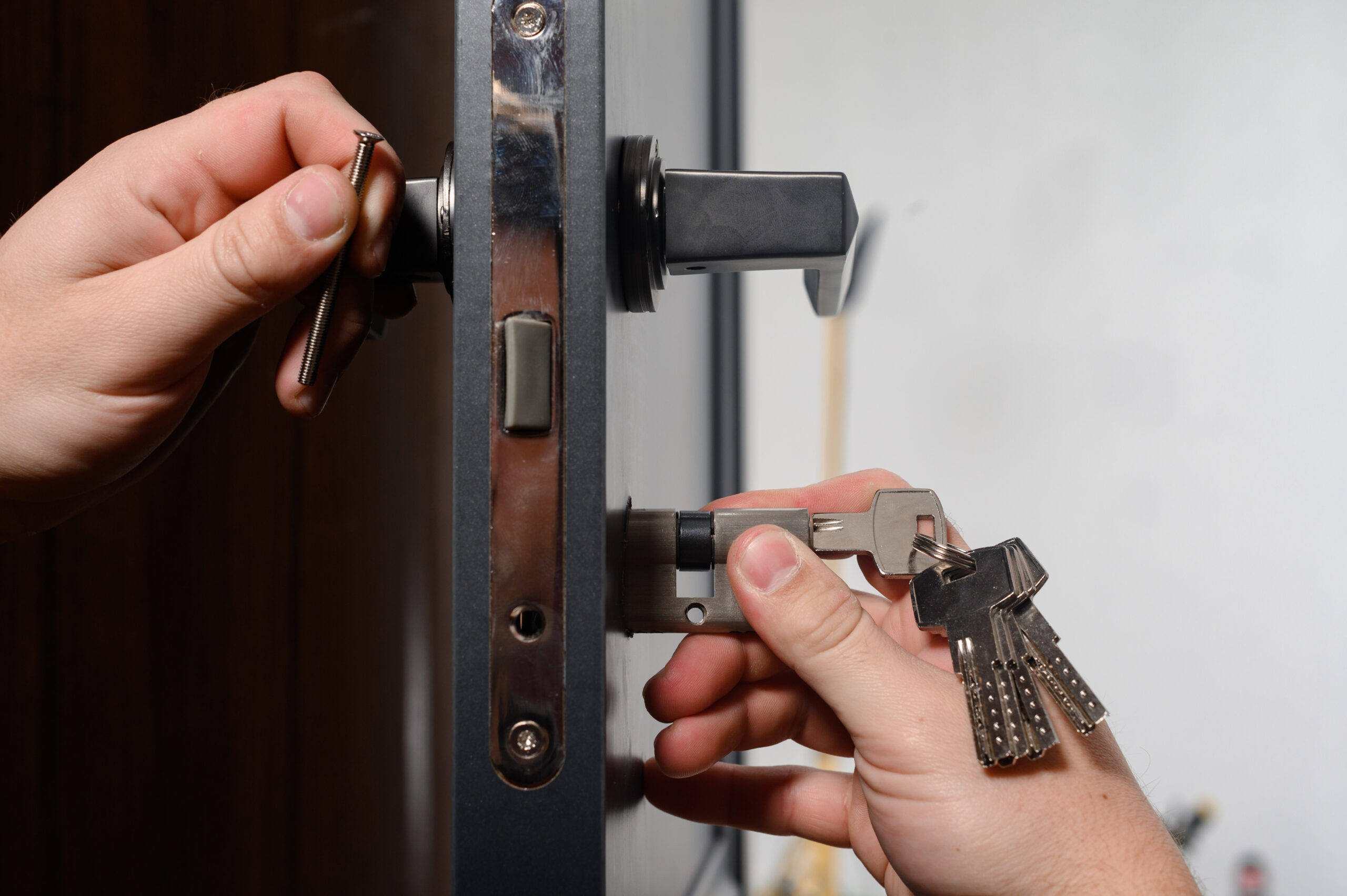 A person’s hands are installing a door lock; one hand holds a screwdriver tightening a screw, while the other hand holds a set of keys inserted into the lock’s cylinder.