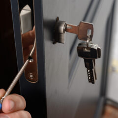 A person uses a screwdriver to repair or install a metal door lock, while a set of keys is inserted into the lock on a blue door.