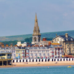 Colorful seaside town with rows of houses, a sandy beach, people walking, and a prominent church with a tall spire in the background. Green hills and blue water complete the coastal scenery.